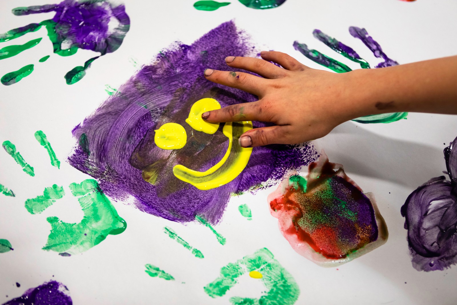 A piece of paper covered in green and purple hand prints. A child's hand draws a yellow smiley face against a purple background in the middle.