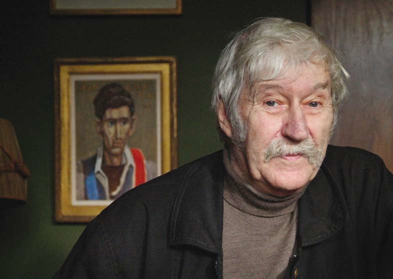John Craxton Photograph of a man with white hair and moustache in front of a portrait of a young man with dark hair.