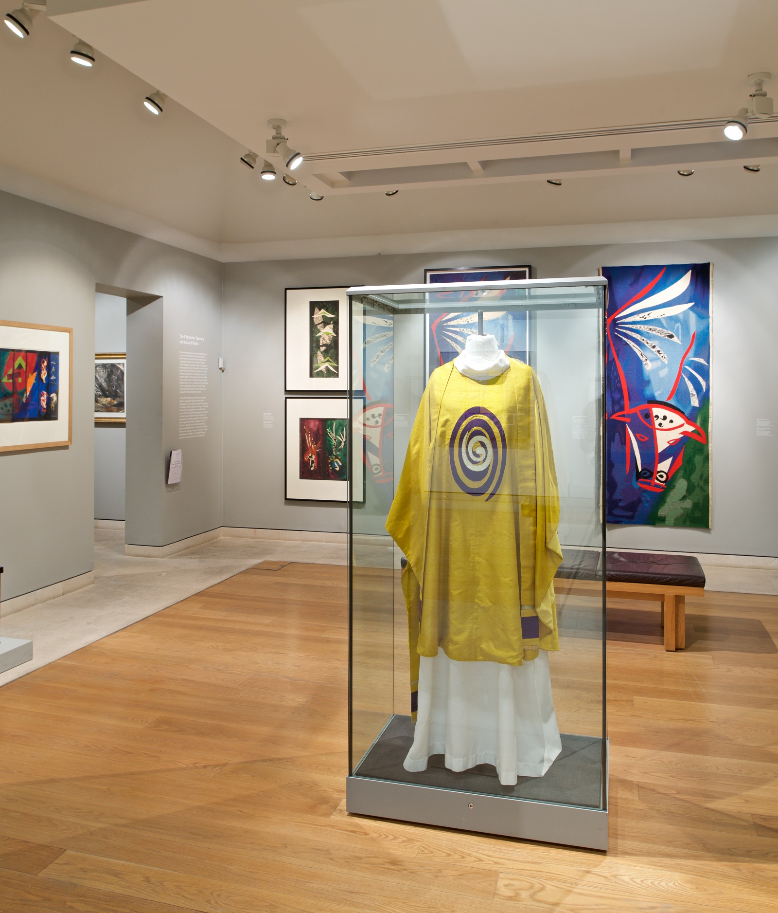Photograph Mark Heathcote View of gallery showing abstract works of art on the wall. In the centre is a glass case containing a mannequin wearing a yellow vestement with abstract oval design on its chest