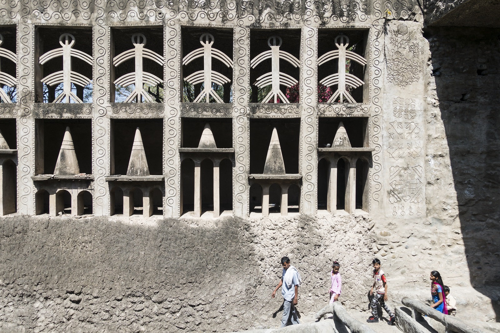 Nek Chand, The Rock Garden of Chandigarh Three men stand beneath a huge concrete wall with recesses filled with abstract sculptures.