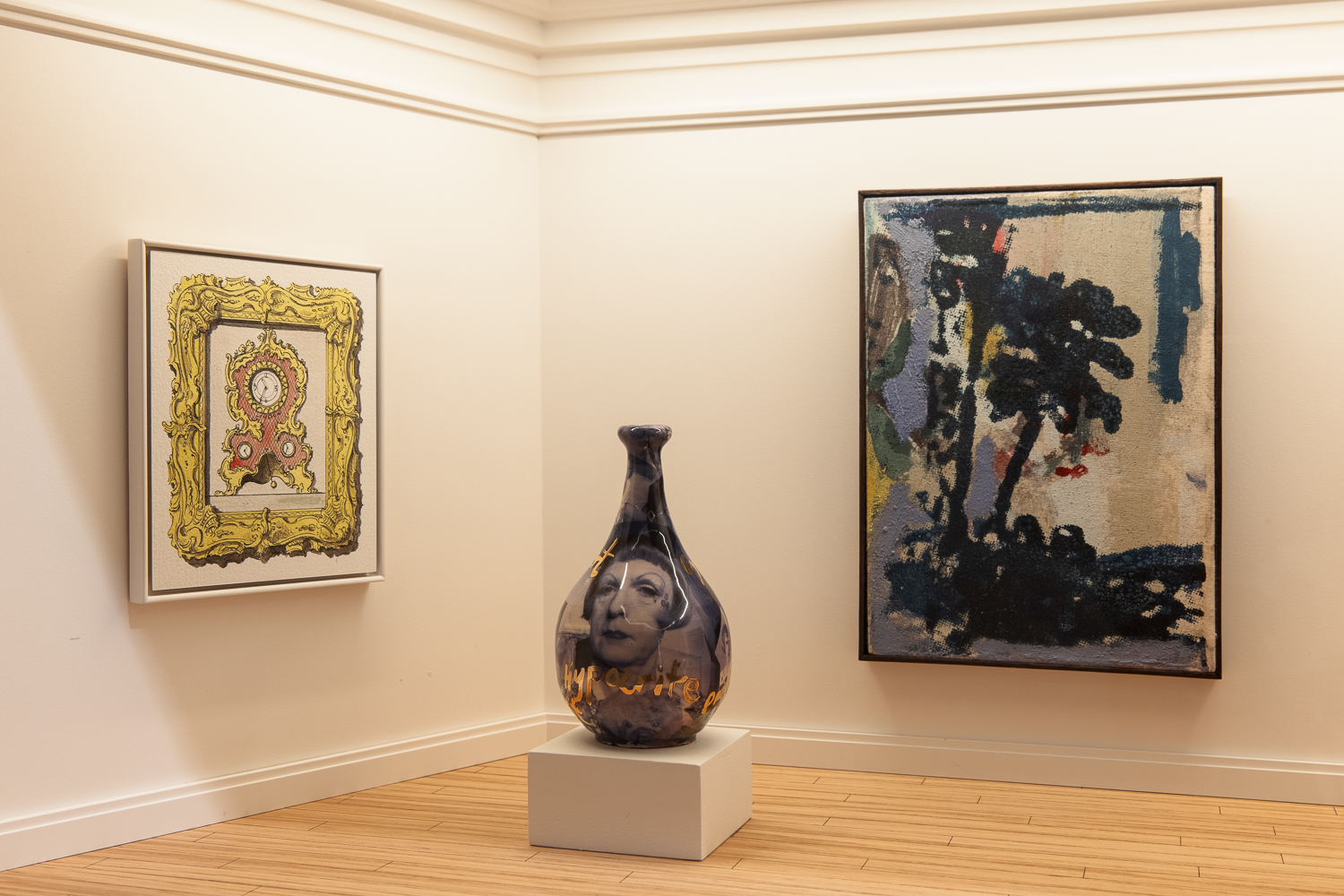 Three works of art in a gallery with white walls and wooden flooring. On the left hand wall is a painting of a baroque clock with an ornate frame. On the right hand wall is an abstract painting in tones of blue, green and white. On a plinth in front of them is a ceramic bottle vase with a picture of the artist Grayson Perry with gold writing.