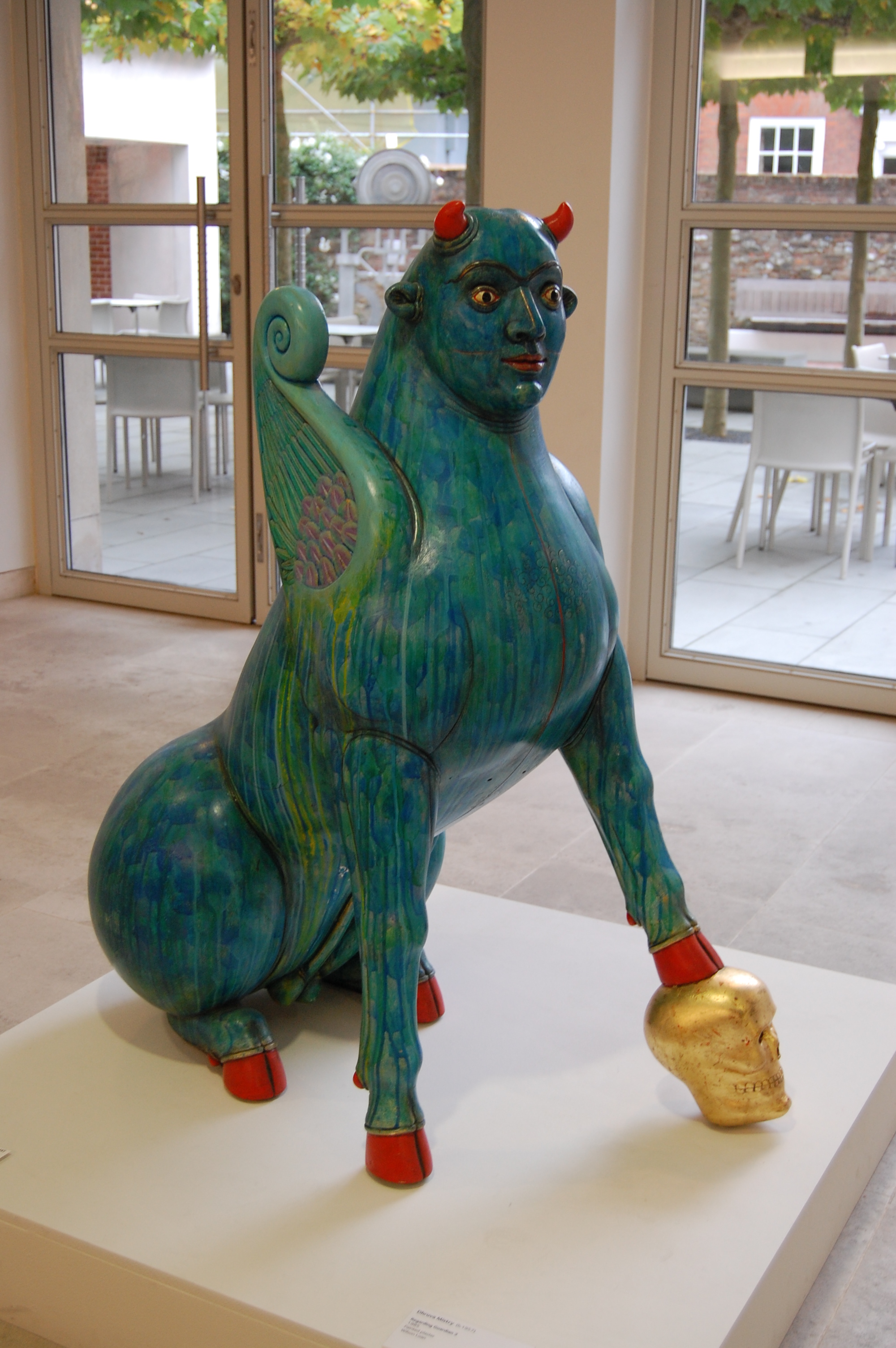 Sculpture of a blue creature with horns and wings and red hooves with one leg standing on gold skull